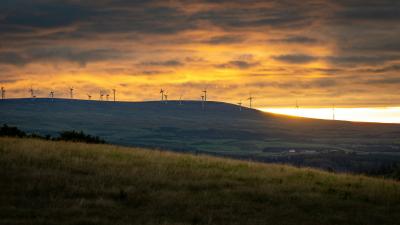 Photo by Carl Jorgensen (https://unsplash.com/@scamartist) on Unsplash (https://unsplash.com/photos/wind-turbines-on-green-grass-field-during-sunset-HOmkYft7Go8) atmospheric sunset over a wind farm with clouds and hill